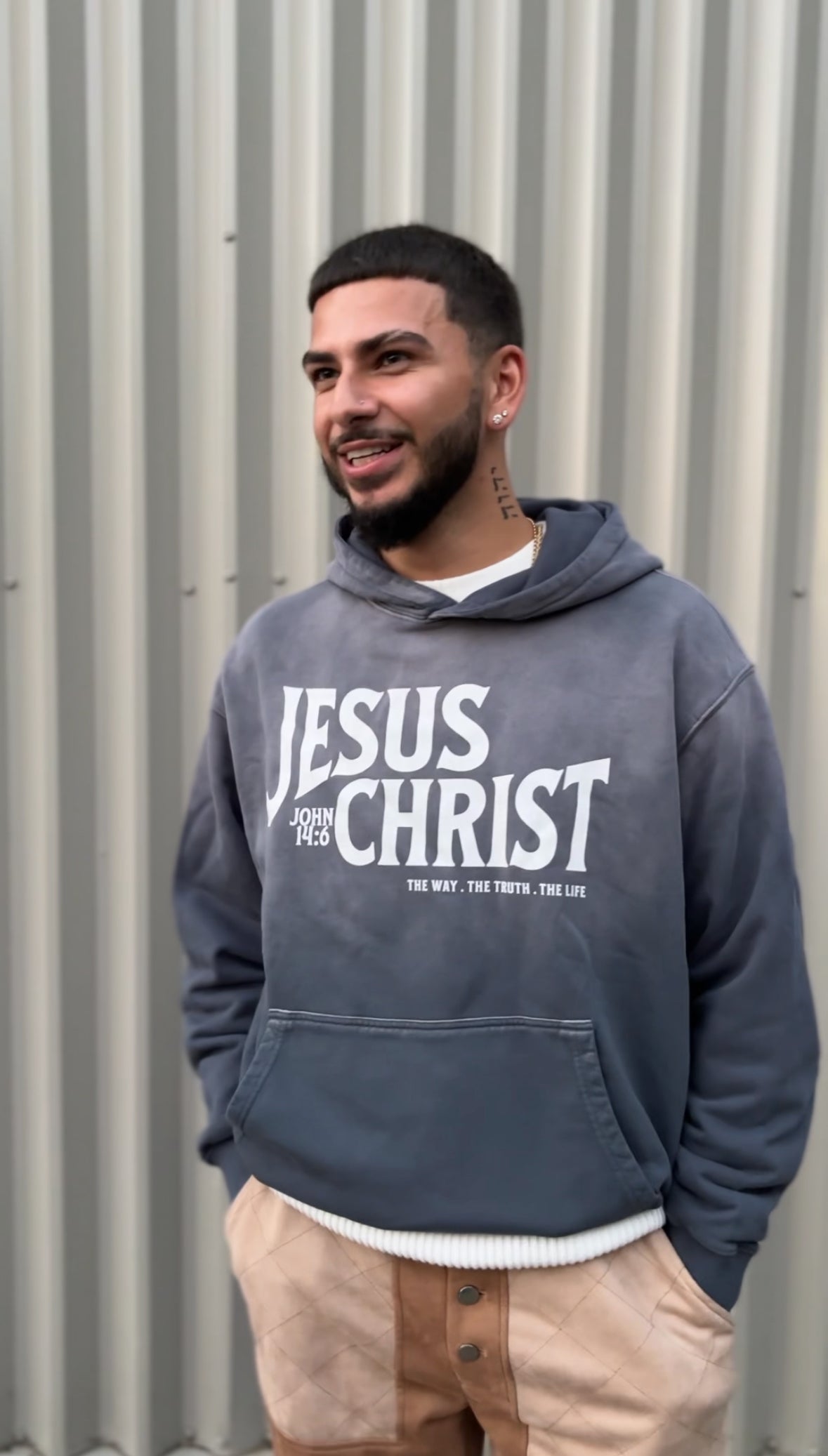 Man wearing a gray hoodie with 'Jesus Christ' text against a corrugated metal background