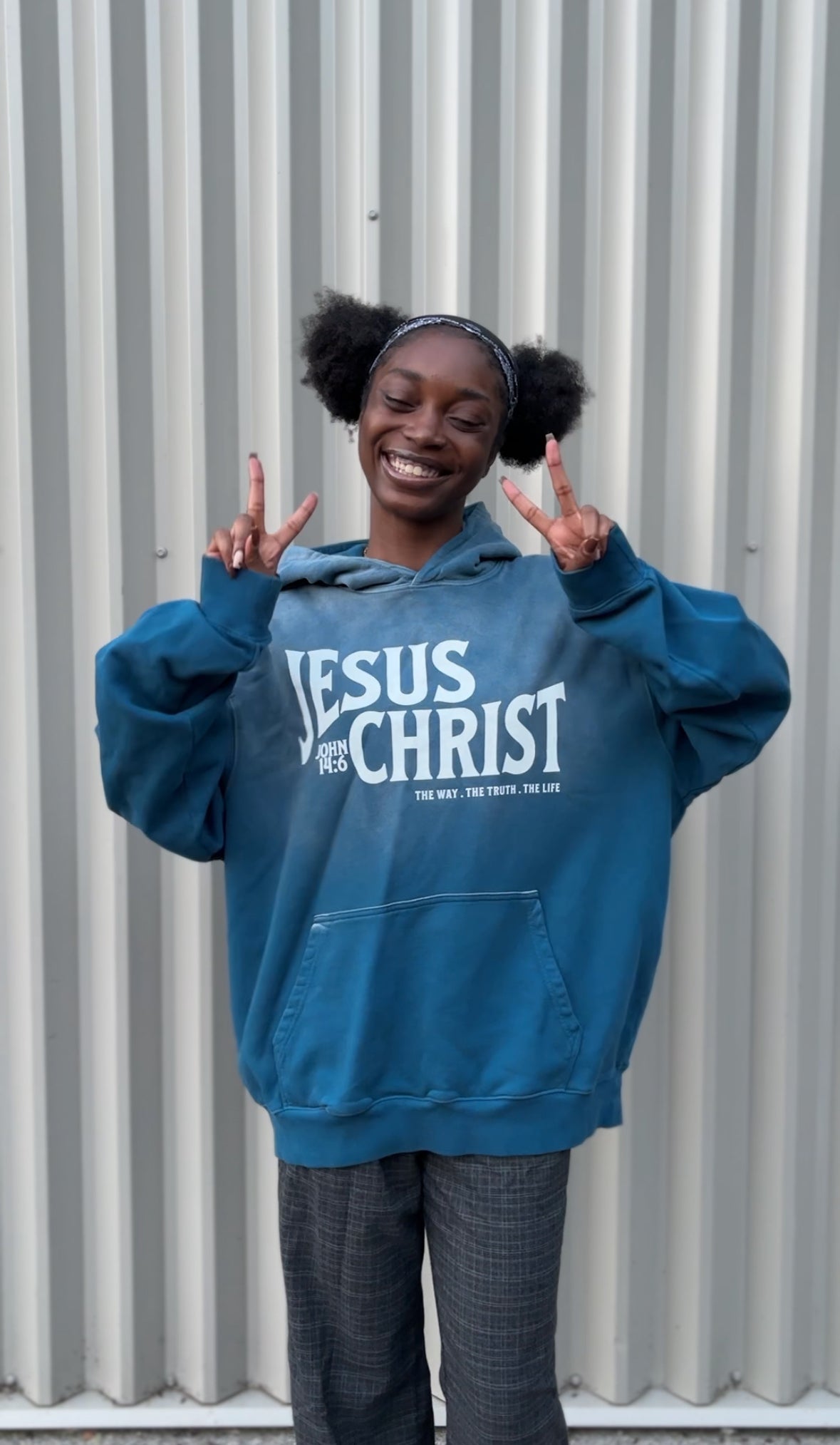 Person wearing a blue hoodie with 'Jesus is the Christ' text, making peace signs in front of a corrugated metal wall.