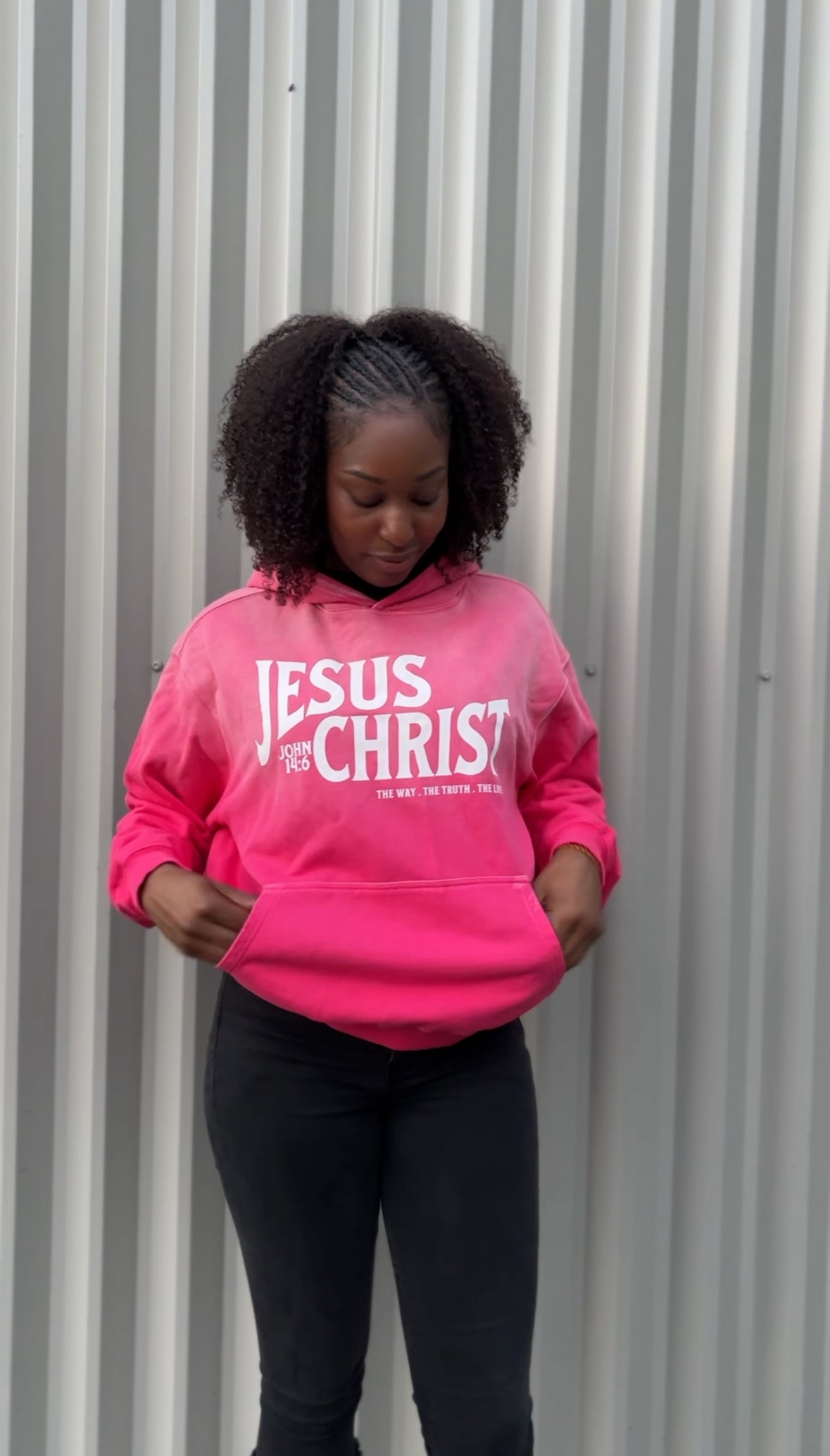 Person wearing a pink hoodie with 'Jesus  Christ' text against a corrugated metal background